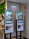 Two digital kiosks for placing takeout orders, displaying images of food items. They are located in a restaurant setting with visible payment interfaces and a sign above indicating 'Takeout Order' in both English and Chinese.
