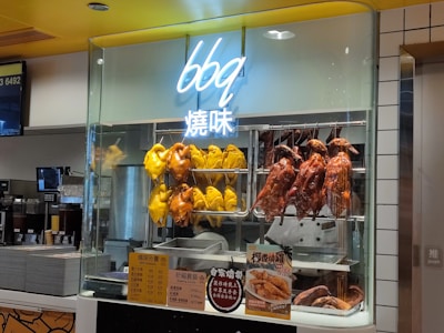 Roasted poultry items, including yellow chickens and brown ducks, are displayed hanging inside a glass case in a food setting. Menu signage and advertisement visuals are visible below the display, with cooking equipment in the background.