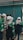 A group of individuals wearing white helmets and green uniform jackets stands near a corrugated metal wall. They appear to be engaged in some activity, possibly an inspection or discussion, holding documents and writing materials. One individual is wearing a headscarf under the helmet.