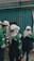 A group of individuals wearing white helmets and green uniform jackets stands near a corrugated metal wall. They appear to be engaged in some activity, possibly an inspection or discussion, holding documents and writing materials. One individual is wearing a headscarf under the helmet.