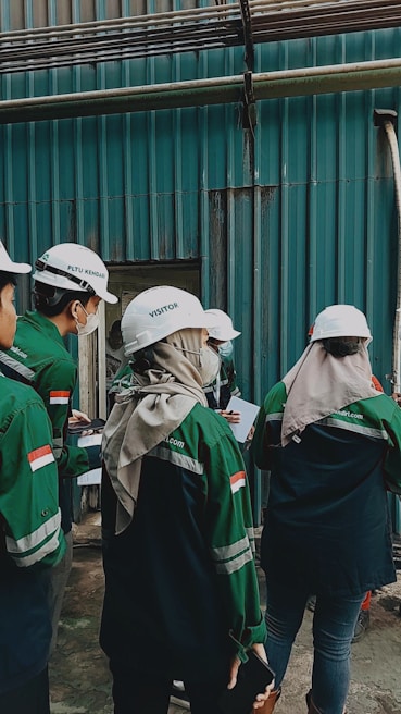 A group of individuals wearing white helmets and green uniform jackets stands near a corrugated metal wall. They appear to be engaged in some activity, possibly an inspection or discussion, holding documents and writing materials. One individual is wearing a headscarf under the helmet.