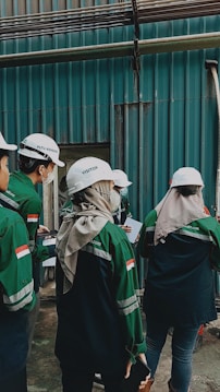 A group of individuals wearing white helmets and green uniform jackets stands near a corrugated metal wall. They appear to be engaged in some activity, possibly an inspection or discussion, holding documents and writing materials. One individual is wearing a headscarf under the helmet.