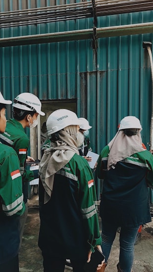 A group of individuals wearing white helmets and green uniform jackets stands near a corrugated metal wall. They appear to be engaged in some activity, possibly an inspection or discussion, holding documents and writing materials. One individual is wearing a headscarf under the helmet.