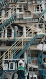 A complex network of industrial stairs, platforms, and pipes with a person wearing a hard hat and safety attire standing at the bottom. The environment is filled with metallic structures and equipment, indicating a manufacturing or industrial setting.