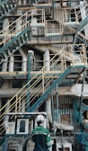 A complex network of industrial stairs, platforms, and pipes with a person wearing a hard hat and safety attire standing at the bottom. The environment is filled with metallic structures and equipment, indicating a manufacturing or industrial setting.