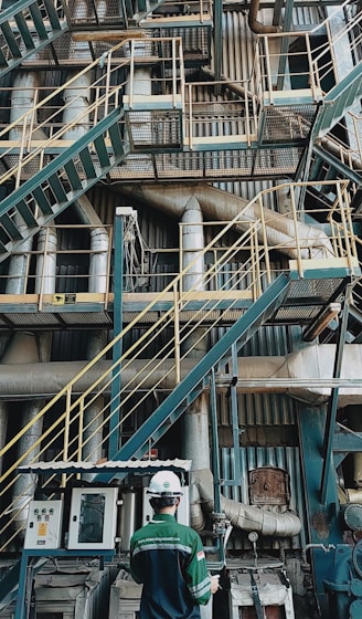 A complex network of industrial stairs, platforms, and pipes with a person wearing a hard hat and safety attire standing at the bottom. The environment is filled with metallic structures and equipment, indicating a manufacturing or industrial setting.