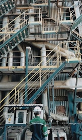 A complex network of industrial stairs, platforms, and pipes with a person wearing a hard hat and safety attire standing at the bottom. The environment is filled with metallic structures and equipment, indicating a manufacturing or industrial setting.