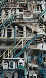 A complex network of industrial stairs, platforms, and pipes with a person wearing a hard hat and safety attire standing at the bottom. The environment is filled with metallic structures and equipment, indicating a manufacturing or industrial setting.