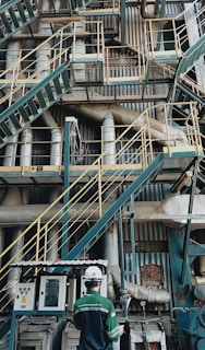 A complex network of industrial stairs, platforms, and pipes with a person wearing a hard hat and safety attire standing at the bottom. The environment is filled with metallic structures and equipment, indicating a manufacturing or industrial setting.