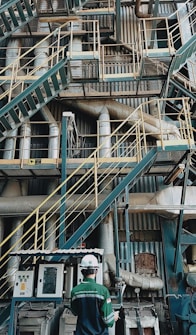 A complex network of industrial stairs, platforms, and pipes with a person wearing a hard hat and safety attire standing at the bottom. The environment is filled with metallic structures and equipment, indicating a manufacturing or industrial setting.