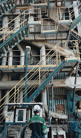 A complex network of industrial stairs, platforms, and pipes with a person wearing a hard hat and safety attire standing at the bottom. The environment is filled with metallic structures and equipment, indicating a manufacturing or industrial setting.