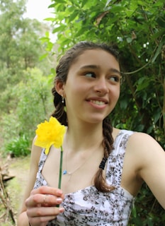A young woman with braided hair is holding a yellow flower in a lush green environment. She is wearing a patterned dress and looking off to the side with a gentle smile. The background is filled with dense foliage.