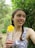 A young woman with braided hair is holding a yellow flower in a lush green environment. She is wearing a patterned dress and looking off to the side with a gentle smile. The background is filled with dense foliage.