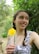 A young woman with braided hair is holding a yellow flower in a lush green environment. She is wearing a patterned dress and looking off to the side with a gentle smile. The background is filled with dense foliage.