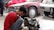 A mechanic wearing a red shirt and working gloves is crouched down, focusing on repairing a car part, likely a transmission. The scene takes place in a garage with multiple cars parked, including a maroon car in the center and a silver car on the right.