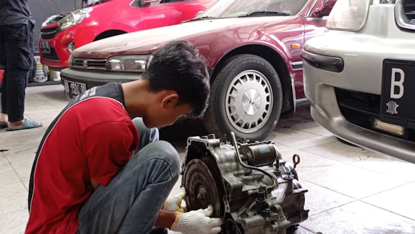A mechanic wearing a red shirt and working gloves is crouched down, focusing on repairing a car part, likely a transmission. The scene takes place in a garage with multiple cars parked, including a maroon car in the center and a silver car on the right.