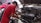A mechanic is working on an open car engine, with visible components including the Honda VTEC engine. The person is wearing a red shirt and white gloves, focused on adjusting or repairing the engine. There are several cars in the background, indicating a workshop environment.