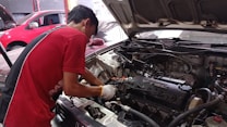 A mechanic is working on an open car engine, with visible components including the Honda VTEC engine. The person is wearing a red shirt and white gloves, focused on adjusting or repairing the engine. There are several cars in the background, indicating a workshop environment.