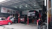 A busy automotive workshop with multiple cars being serviced. One car is elevated on a lift, and a mechanic is working nearby. Several signs and banners with automotive-related text hang on the walls. The environment is industrial, with metal roofing and visible beams.