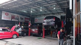 A busy automotive workshop with multiple cars being serviced. One car is elevated on a lift, and a mechanic is working nearby. Several signs and banners with automotive-related text hang on the walls. The environment is industrial, with metal roofing and visible beams.