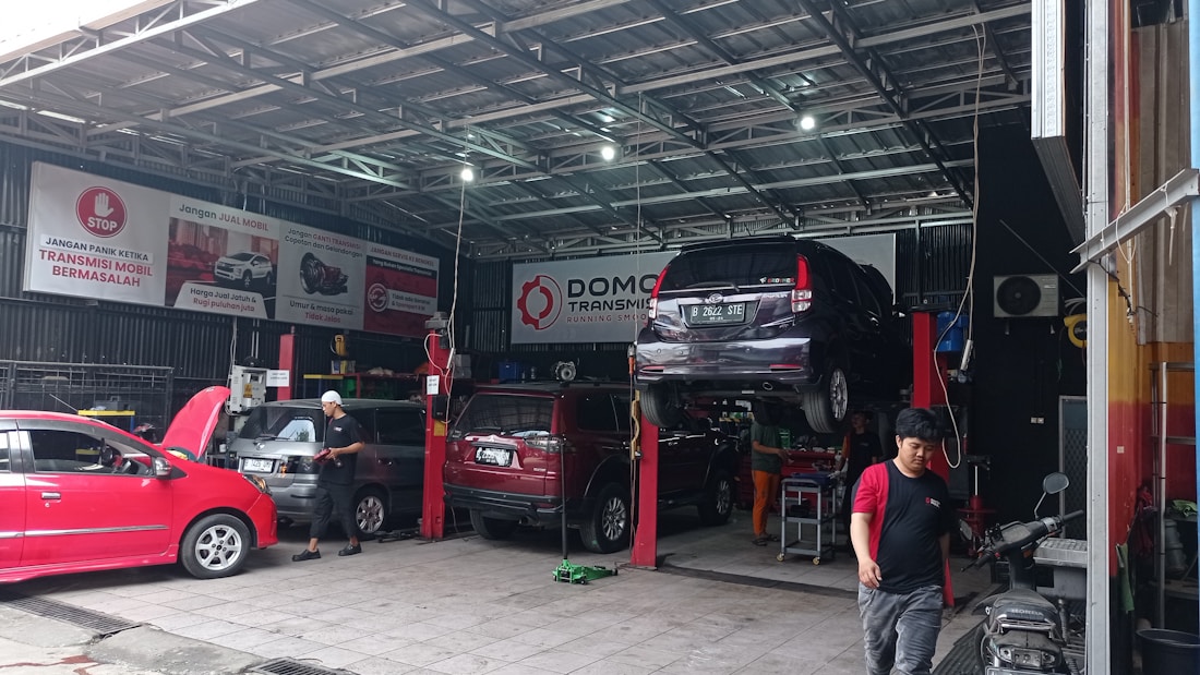 A busy automotive workshop with multiple cars being serviced. One car is elevated on a lift, and a mechanic is working nearby. Several signs and banners with automotive-related text hang on the walls. The environment is industrial, with metal roofing and visible beams.