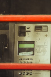 The image depicts a close-up view of a public payphone panel, featuring a numeric keypad, coin return slot, and instruction labels. The panel seems to be behind a red frame, possibly part of a traditional phone booth. The display screen is present above the numeric pad.