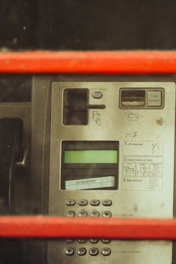 The image depicts a close-up view of a public payphone panel, featuring a numeric keypad, coin return slot, and instruction labels. The panel seems to be behind a red frame, possibly part of a traditional phone booth. The display screen is present above the numeric pad.