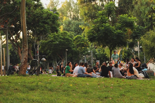 A group of people sitting on the grass in a park, engaging in conversation and enjoying an outdoor gathering. Trees create a shaded canopy above, and additional people can be seen in the background walking and biking. A table with refreshments is nearby.