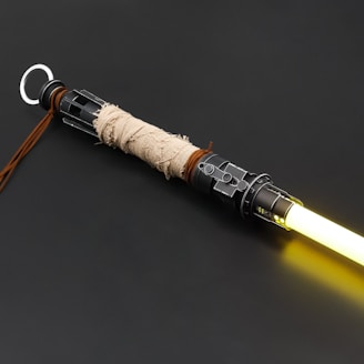 A close-up of a lightsaber hilt with intricate details, including a weathered fabric wrap and metallic parts. The lightsaber emits a bright yellow glow from its blade, and a leather strap hangs from the end of the hilt.