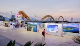 An outdoor exhibition with large photo panels and a sign that reads 'Da Nang' under a dragon-shaped bridge. A person with a backpack stands viewing the display. The background shows a city skyline with tall buildings and mountains under a dusk sky.