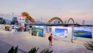 An outdoor exhibition with large photo panels and a sign that reads 'Da Nang' under a dragon-shaped bridge. A person with a backpack stands viewing the display. The background shows a city skyline with tall buildings and mountains under a dusk sky.
