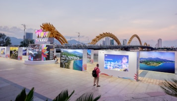 An outdoor exhibition with large photo panels and a sign that reads 'Da Nang' under a dragon-shaped bridge. A person with a backpack stands viewing the display. The background shows a city skyline with tall buildings and mountains under a dusk sky.