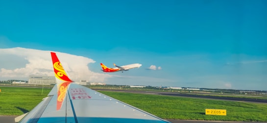 An airplane is taking off against a backdrop of clear blue sky and scattered clouds, as seen from another airplane waiting on the runway. The vibrant colors on the plane's tail stand out against the sky.