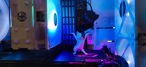 A lit-up computer case with multiple cooling fans and LED lights features a small, white figurine of a cat standing in a playful pose near the center.