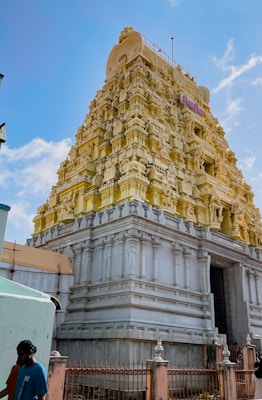 An ornately carved temple tower, or gopuram, characterized by intricate sculptures and a blend of yellow and white hues. The architectural design features multiple tiers of detailed mythology-inspired statues. A clear blue sky serves as the backdrop, enhancing the temple's vibrant colors. In the foreground, a few people are walking past, one of whom is wearing a blue shirt.