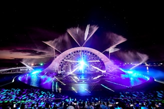 A nighttime water and light show features an illuminated stage with a large, arching structure. The stage is surrounded by water with fountains spraying into the air, lit by vibrant lights in shades of blue and purple. The lights create reflections and patterns on the water's surface, while a crowd of people watches the show from seating in the foreground.