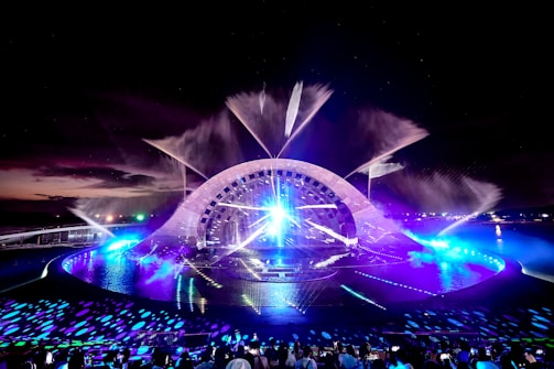 A nighttime water and light show features an illuminated stage with a large, arching structure. The stage is surrounded by water with fountains spraying into the air, lit by vibrant lights in shades of blue and purple. The lights create reflections and patterns on the water's surface, while a crowd of people watches the show from seating in the foreground.