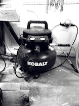 A black Kobalt air compressor is situated on a workshop floor, surrounded by various tools and wooden planks. Hoses are connected to the compressor, and wrenches are hanging on a pegboard above.