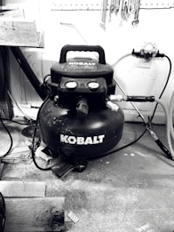 A black Kobalt air compressor is situated on a workshop floor, surrounded by various tools and wooden planks. Hoses are connected to the compressor, and wrenches are hanging on a pegboard above.