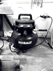A black Kobalt air compressor is situated on a workshop floor, surrounded by various tools and wooden planks. Hoses are connected to the compressor, and wrenches are hanging on a pegboard above.
