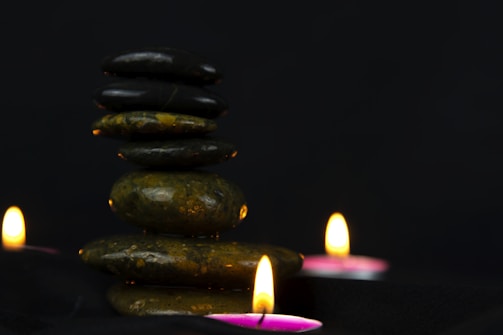 A stack of smooth, rounded stones is balanced in the center against a dark background. Three small tealight candles with gentle flames are positioned around the stones, casting a warm glow.