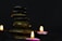 A stack of smooth, rounded stones is balanced in the center against a dark background. Three small tealight candles with gentle flames are positioned around the stones, casting a warm glow.