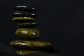 A stack of smooth, polished stones is arranged in a balance against a dark background. The stones vary in size and color, featuring shades of black, green, and brown. The arrangement suggests calmness and stability, with each stone carefully placed on top of the other.