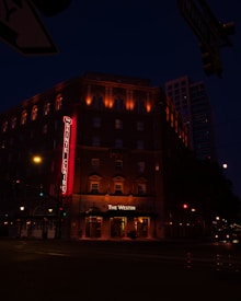 A nighttime urban street scene featuring a multi-story building with illuminated signs and lights. The building, likely a hotel, displays the names 'The Sainte Claire' and 'The Westin' in bright lights. The streets are relatively empty, with some streetlights and traffic signals visible.