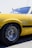 A close-up view of a bright yellow classic car, focusing on the side panel and front wheel. The wheel is fitted with a chrome hubcap and black radial tires, and there are some decals and insignia on the car's side. The vehicle appears to be parked outdoors with a clear blue sky in the background.