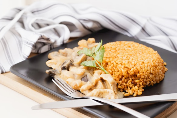 On a black square plate, there is a serving of golden brown cooked grains shaped in a dome next to a creamy mushroom sauce. Fresh herbs garnish the dish. A silver fork and knife are placed on the edge of the plate, and a black and white striped cloth is draped in the background.