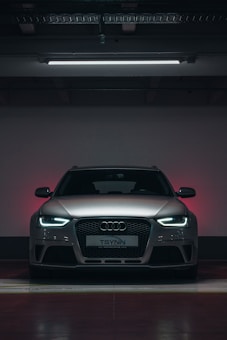 A silver Audi car is parked in a dimly lit indoor parking space. The car's headlights are turned on with bright white light, and the front grille and logo are prominently visible. The background has a subtle red and grey gradient.