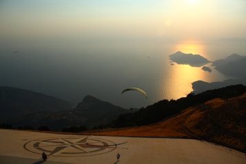 A scenic coastal landscape viewed from a high vantage point. The sun sets over the calm sea, casting a golden reflection across the water. Several paragliders soar in the sky, with one prominently in the foreground. Rugged terrain and small islands are visible in the distance, creating a serene and expansive atmosphere.