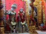 Two intricately designed statues are adorned with colorful garlands and traditional Indian attire. The figures stand amidst ornate golden pillars decorated with floral motifs. The background features bright red drapery and additional decorative elements.