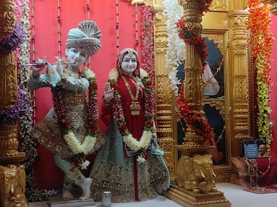 Two intricately designed statues are adorned with colorful garlands and traditional Indian attire. The figures stand amidst ornate golden pillars decorated with floral motifs. The background features bright red drapery and additional decorative elements.